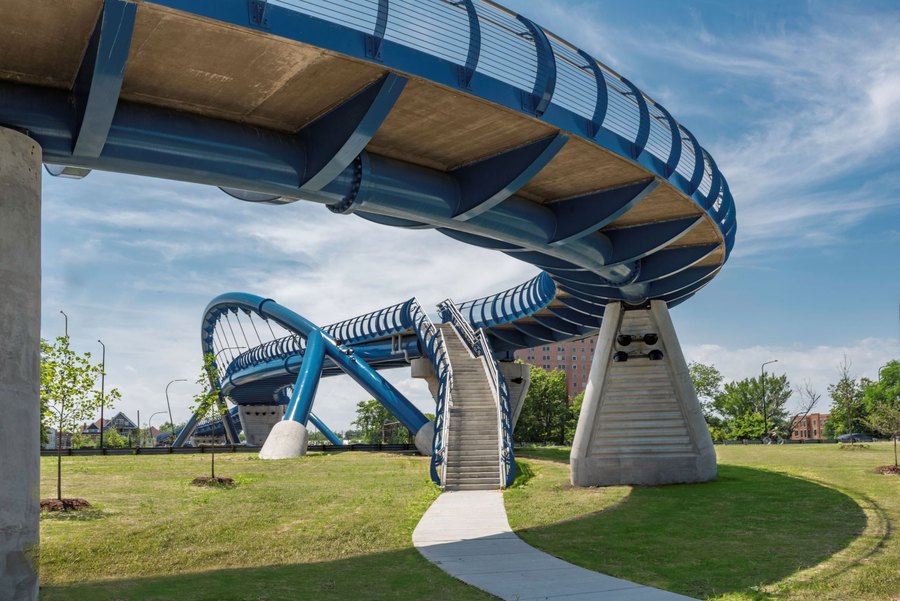 A curving blue steel pedestrian bridge over a landscaped park.