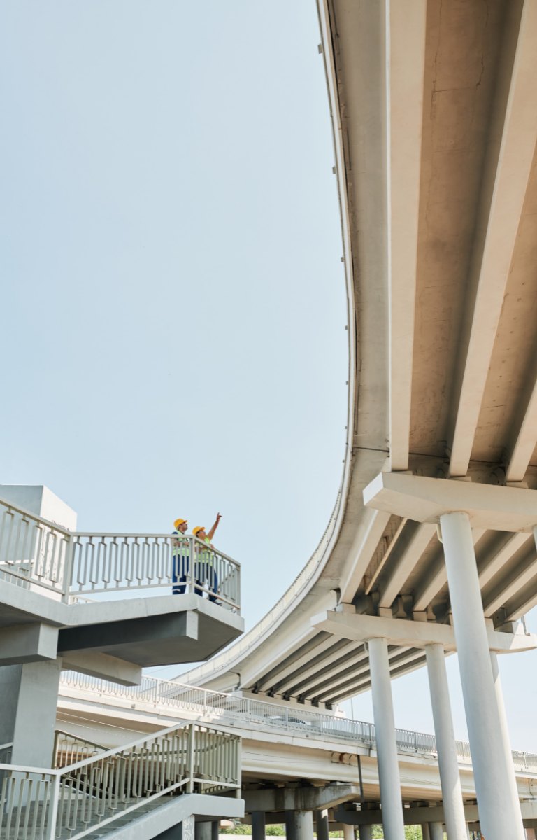 Two HBM engineers in high-visibility gear inspecting an elevated highway interchange